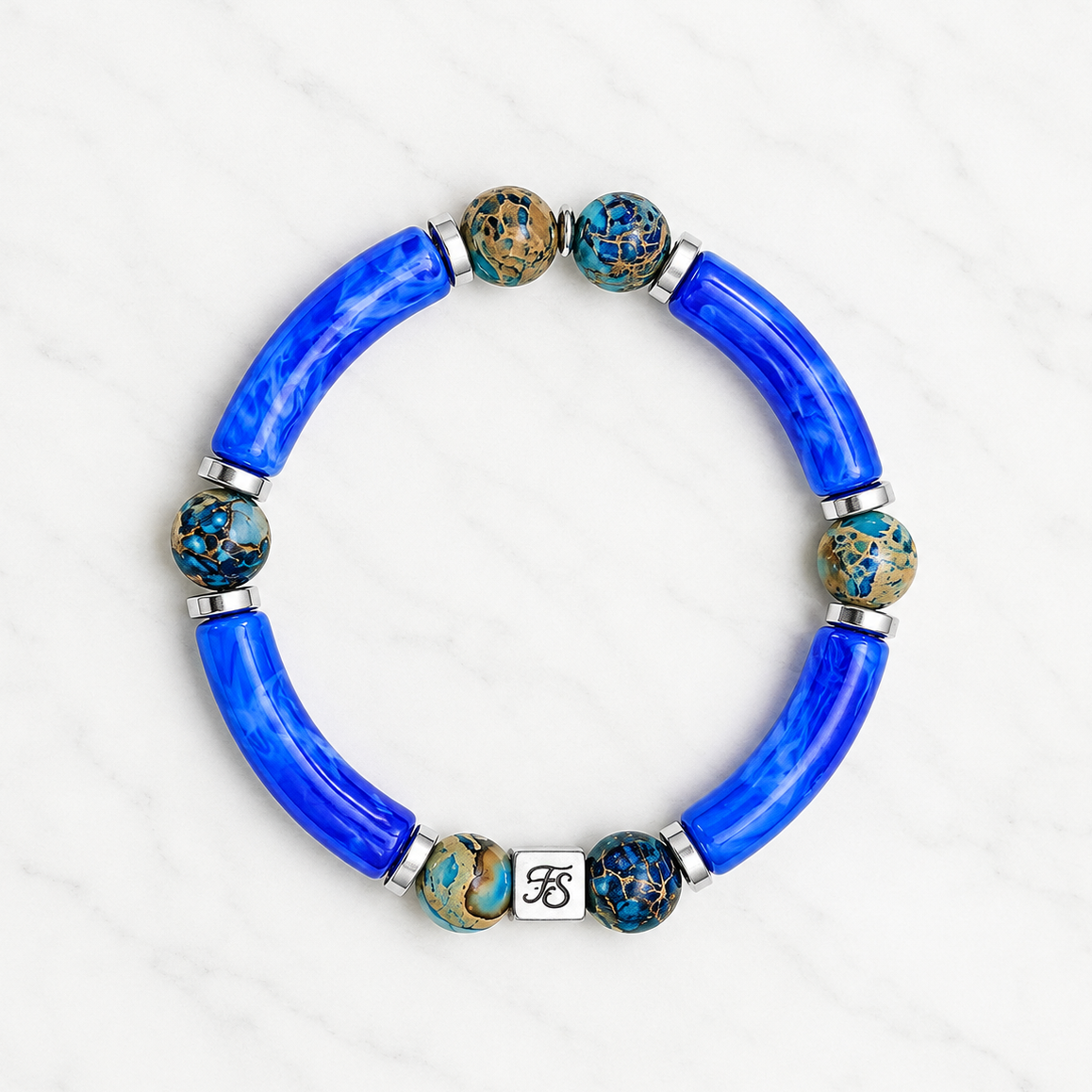 Atlantic Cove Bracelet featuring Blue Imperial Sea Sediment Jasper gemstone beads, cobalt blue acrylic tube beads, polished silver spacer beads and a stainless steel Fleur & Stone logo bead on a light white marble background.