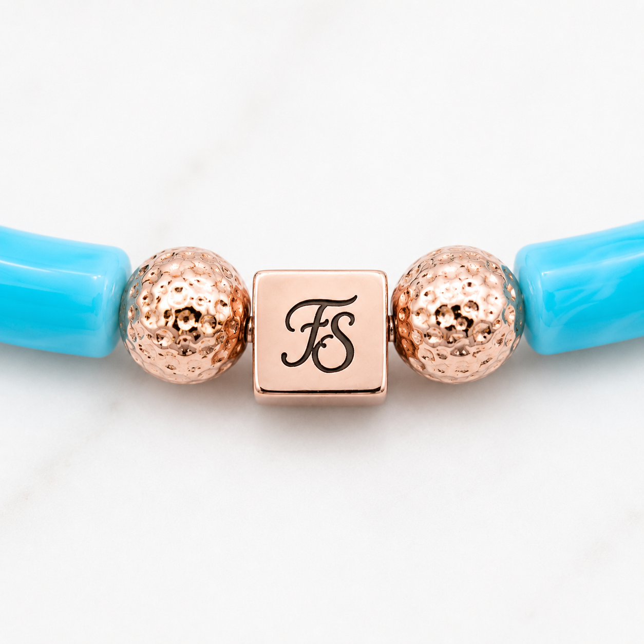Close-up of the Fleur & Stone rose gold stainless steel logo bead with textured rose gold lava beads and turquoise acrylic tube beads on the Pink Lagoon Bracelet.