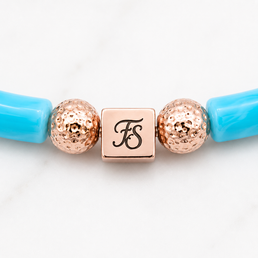 Close-up of the Fleur & Stone rose gold stainless steel logo bead with textured rose gold lava beads and turquoise acrylic tube beads on the Pink Lagoon Bracelet.