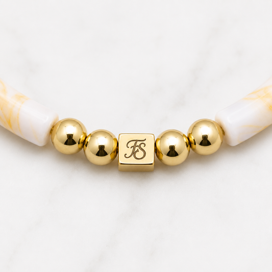 Close-up of the Fleur & Stone gold stainless steel logo bead with polished gold hematite beads and ivory marbled acrylic tube beads on the Golden Sands Bracelet.
