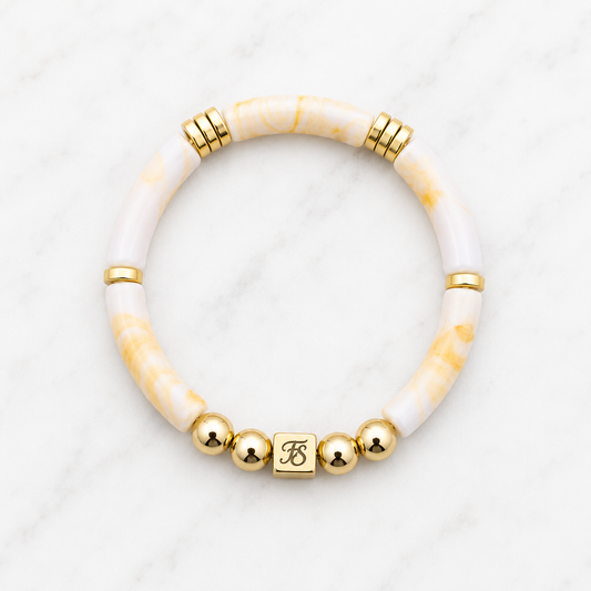 Golden Sands Bracelet by Fleur & Stone featuring ivory and golden marbled acrylic tube beads, polished gold hematite beads and a gold stainless steel logo bead on a white marble background.