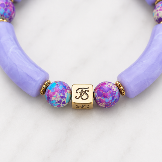Close-up of the Fleur & Stone gold stainless steel logo bead with polished gold spacer beads, Purple Galaxy Sea Sediment Jasper gemstone beads and lavender acrylic tube beads on the Galaxy Skies Bracelet.