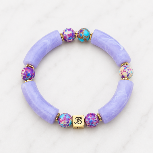 Galaxy Skies Bracelet by Fleur & Stone featuring lavender acrylic tube beads, Purple Galaxy Sea Sediment Jasper gemstone beads, polished gold spacer beads and a gold stainless steel logo bead on a white marble background.