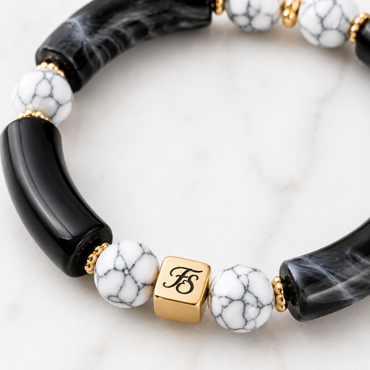 Close-up of the Midnight Marble bracelet showing the gold Fleur & Stone logo bead, white howlite gemstone beads, gold hematite spacer beads, and glossy black marbled acrylic tube beads.
